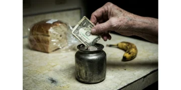 A hand placing a worn dollar bill into a tarnished coin jar, symbolizing the erosion of purchasing power due to economic trends.