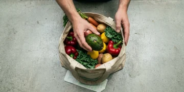 A person's hands carefully placing an avocado into a reusable shopping bag filled with fresh produce, on a concrete counter with a grocery receipt, illustrating the impact of economic shifts on household budgets.