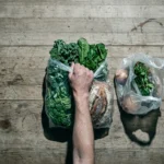 Overhead view of a person's hand holding a full grocery bag next to a visibly smaller, less full bag, symbolizing inflation and shrinkflation eroding purchasing power on a weathered kitchen table.
