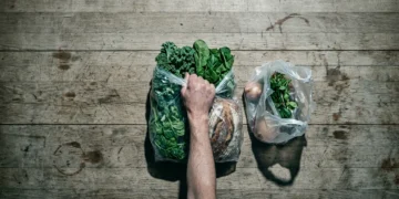 Overhead view of a person's hand holding a full grocery bag next to a visibly smaller, less full bag, symbolizing inflation and shrinkflation eroding purchasing power on a weathered kitchen table.